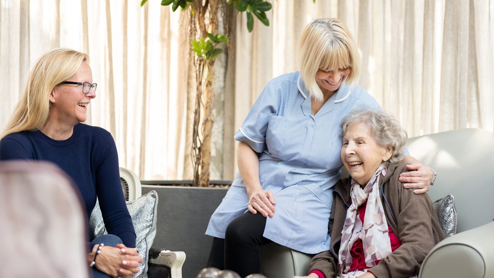 A carer with her arm around a smiling resident, with a visiting relative beside them